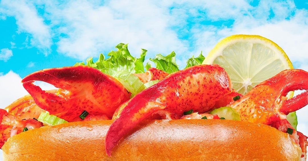 Close-up of a lobster roll with lemon and lettuce against a bright blue sky with white clouds. Food photography, product shot.