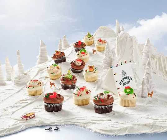 Festive holiday cupcakes arranged on a snowy mountain with paper Christmas trees and a Magnolia Bakery advertisement card.