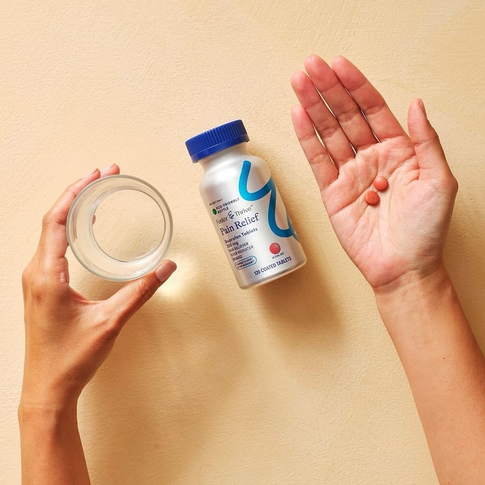 Hands holding a glass of water, pain relief medicine bottle, and two pills against a peach background in a studio setting.