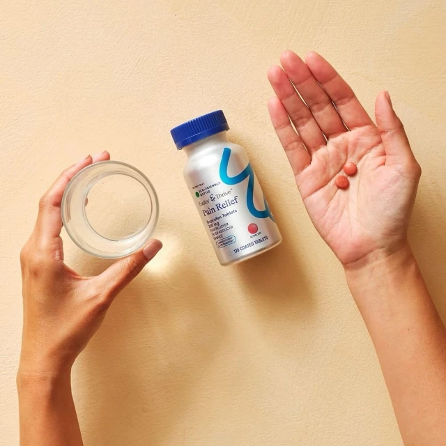 Hands holding a glass of water, pain relief medicine bottle, and two pills against a peach background in a studio setting.