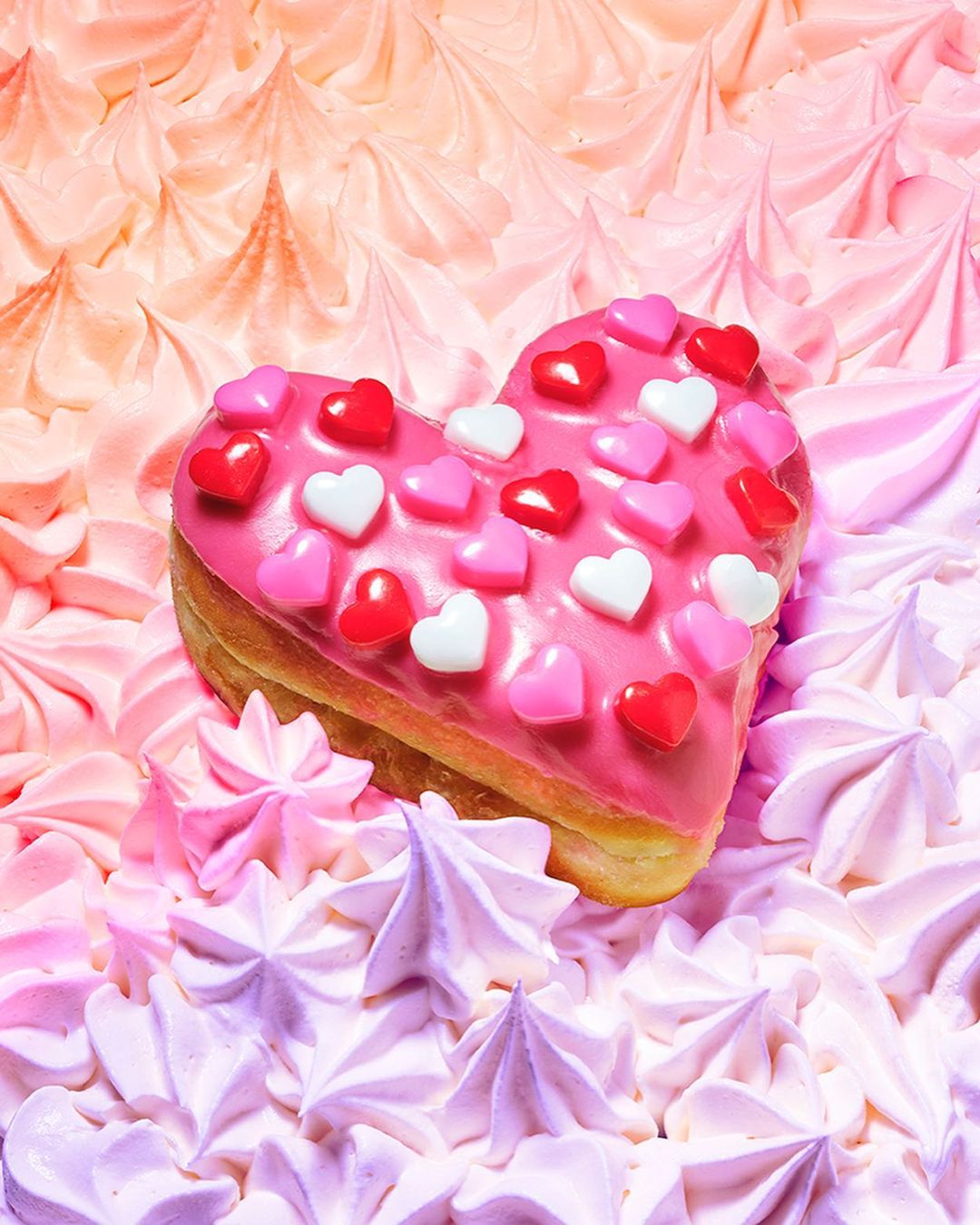 Heart-shaped donut frosted with pink icing and candy hearts, resting on a bed of pink and lavender meringue kisses.