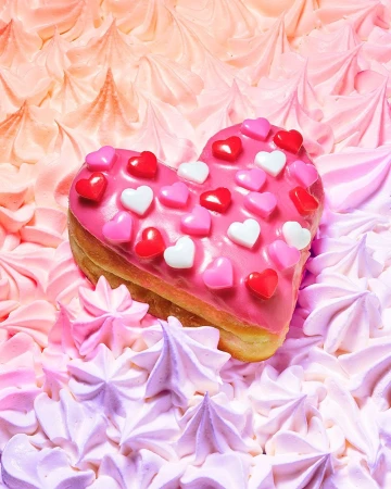 Heart-shaped donut frosted with pink icing and candy hearts, resting on a bed of pink and lavender meringue kisses.