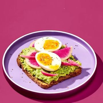 Overhead view of an avocado toast with a sliced soft-boiled egg, pink radishes, and fresh herbs on a purple plate against a pink backdrop.