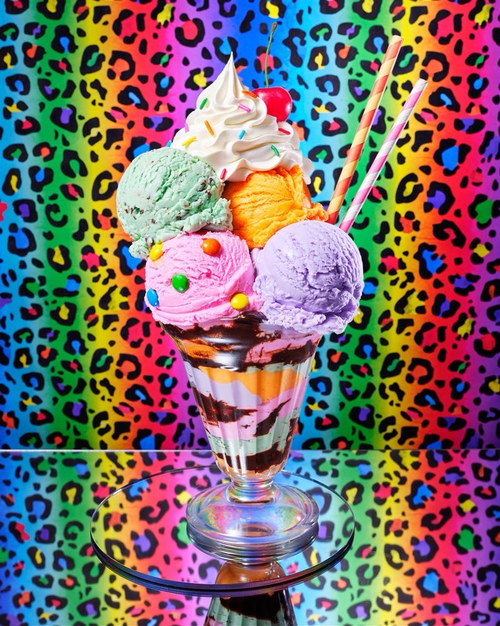 Eye-level studio shot of an ice cream sundae with multiple scoops in a tall glass on a reflective surface against a colorful background.