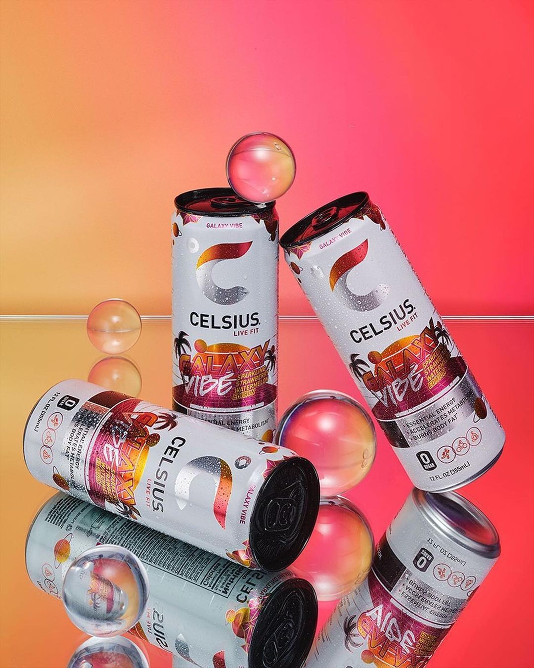 Three cans of Celsius Galaxy Vibe energy drink are featured with clear spheres on a glossy orange to pink gradient surface.