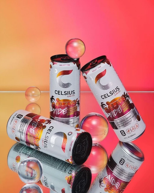 Three cans of Celsius Galaxy Vibe energy drink are featured with clear spheres on a glossy orange to pink gradient surface.