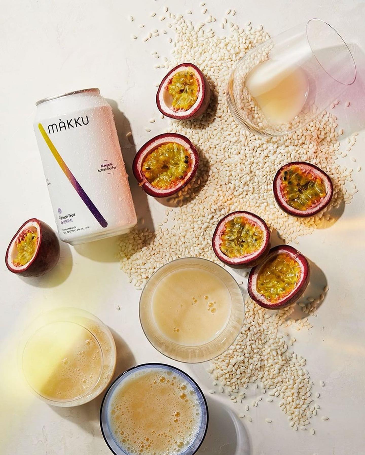 Makkuli rice beverage can with passion fruit halves and glasses of drink surrounded by white grains on a light background. 