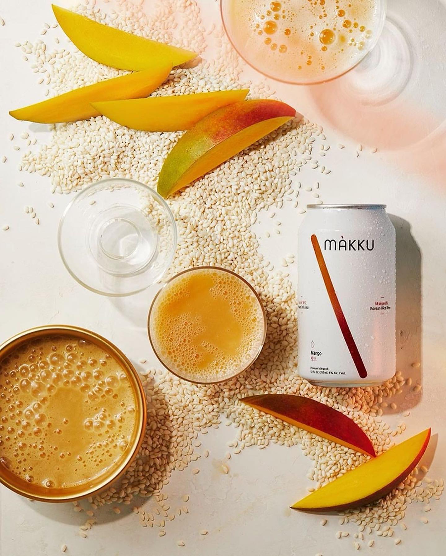 Overhead shot of makku rice beer can and glasses filled with golden liquid and mango slices scattered on white background.