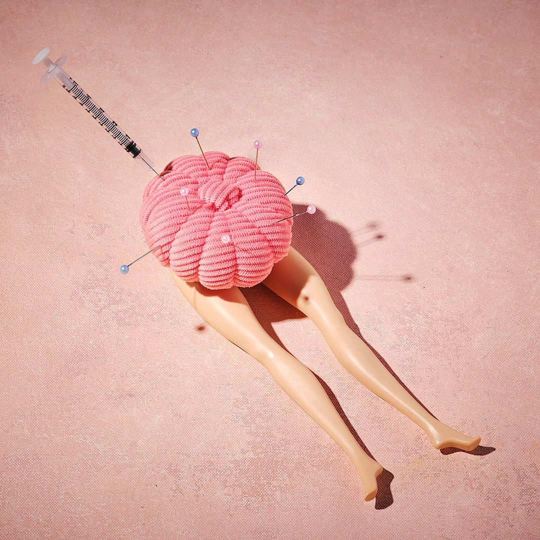 A pink pincushion in the shape of a brain sits atop doll legs, with a syringe entering the brain, all against a pink backdrop.