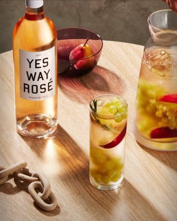 Bottle of rose wine, a glass filled with grapes and beverage, and a decanter on a wood table create an inviting beverage scene.