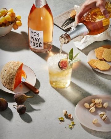 Overhead shot of a hand pouring rosé into a cocktail with a fig, alongside cheese, crackers, grapes, and pistachios on a gray surface.