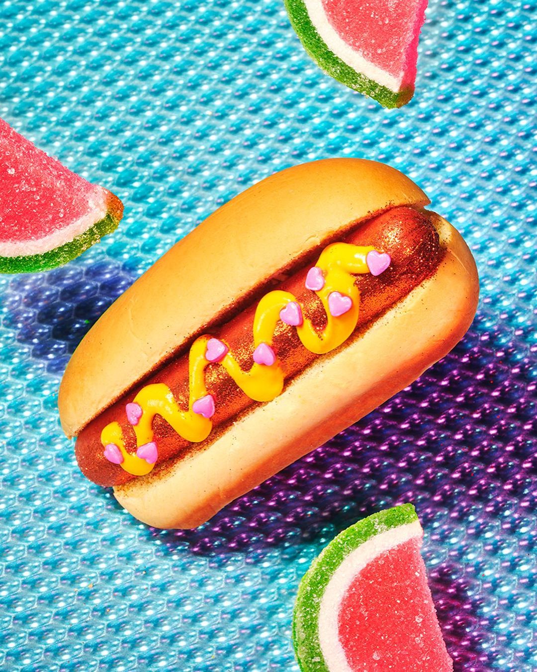 Close-up of a hot dog with mustard and candy hearts, with watermelon candies on a blue bubble-textured background. 