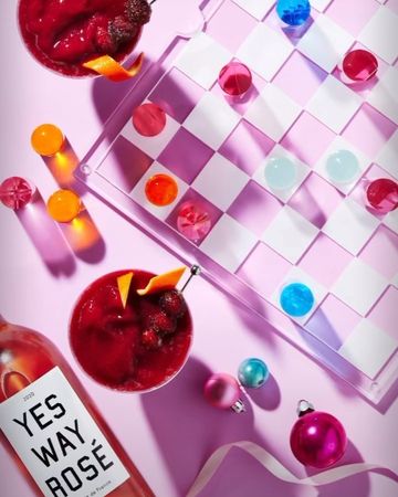 Overhead shot of two berry cocktails, a checkerboard with glass marbles, a bottle of rosé wine and a ribbon on a pink background.