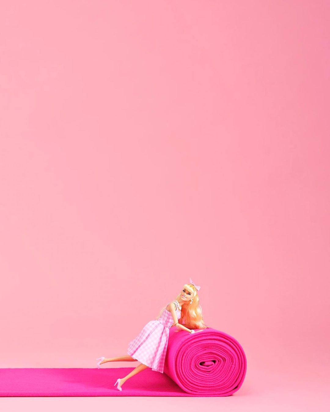 Blonde doll in a pink dress on a pink rolled carpet and a matching pink background for a minimalist, toy-like, artificial aesthetic.