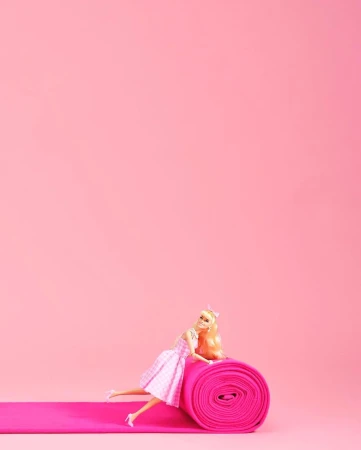 Blonde doll in a pink dress on a pink rolled carpet and a matching pink background for a minimalist, toy-like, artificial aesthetic.