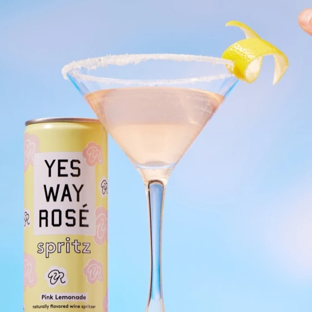 A can of Yes Way Rosé Spritz sits next to a martini glass filled with a pink drink, rimmed with sugar and garnished with a lemon twist.