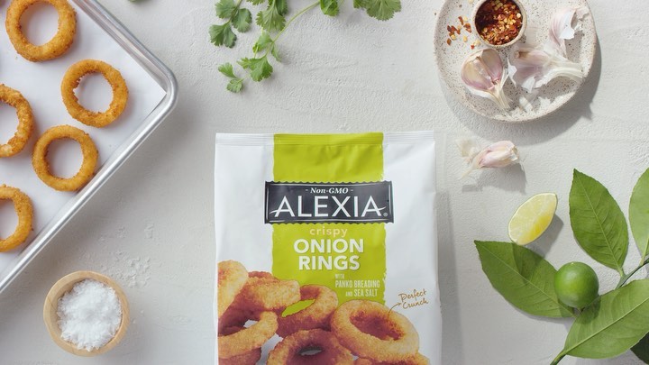 Overhead shot of frozen Alexia onion rings, alongside cilantro, limes, spices, and garlic on a white background, for a food and beverage theme.