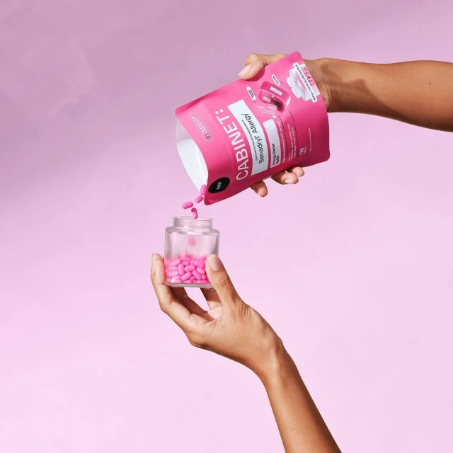 Hands pour pink pills from a pink bag into a clear jar against a solid pink backdrop, showcasing a health and wellness product.