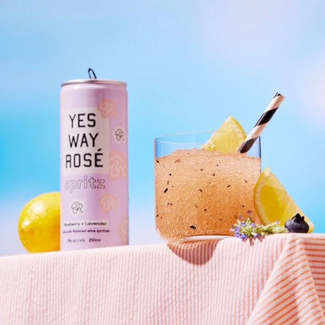 A can of rose spritz and a glass of the same beverage next to a lemon on a table with a striped tablecloth against a blue background.