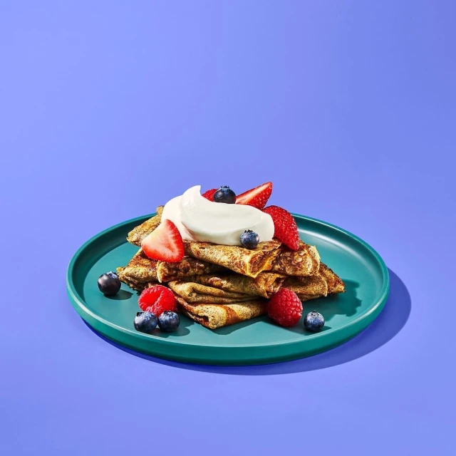 Crepes stacked on a green plate, topped with whipped cream and mixed fresh berries on a violet background in a studio setting.