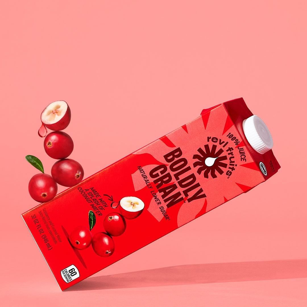 Red cranberry juice box floating in a pink studio setting with cranberries arranged in a vertical stack. Food and beverage still life.