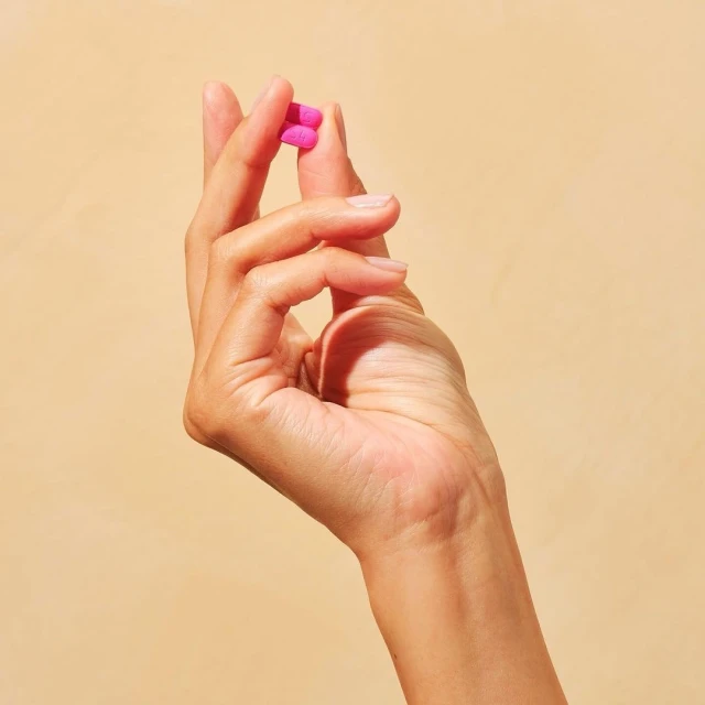 Close-up of a hand holding two pink pills against a tan background, showcasing the product in a simple, uncluttered studio setting.
