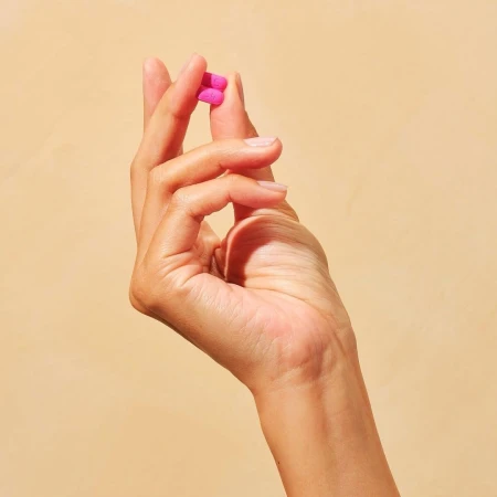 Close-up of a hand holding two pink pills against a tan background, showcasing the product in a simple, uncluttered studio setting.