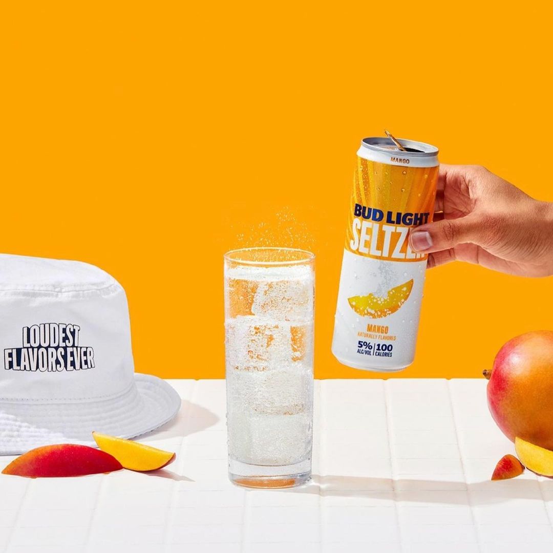 Bud Light Seltzer Mango with a glass of seltzer over a white table, set against an orange background, featuring a white bucket hat.