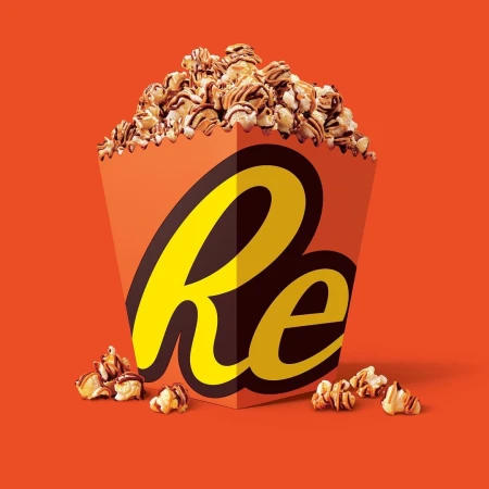Box of Reese's popcorn featuring chocolate and peanut butter drizzle. The popcorn is overflowing from the branded container on an orange background.