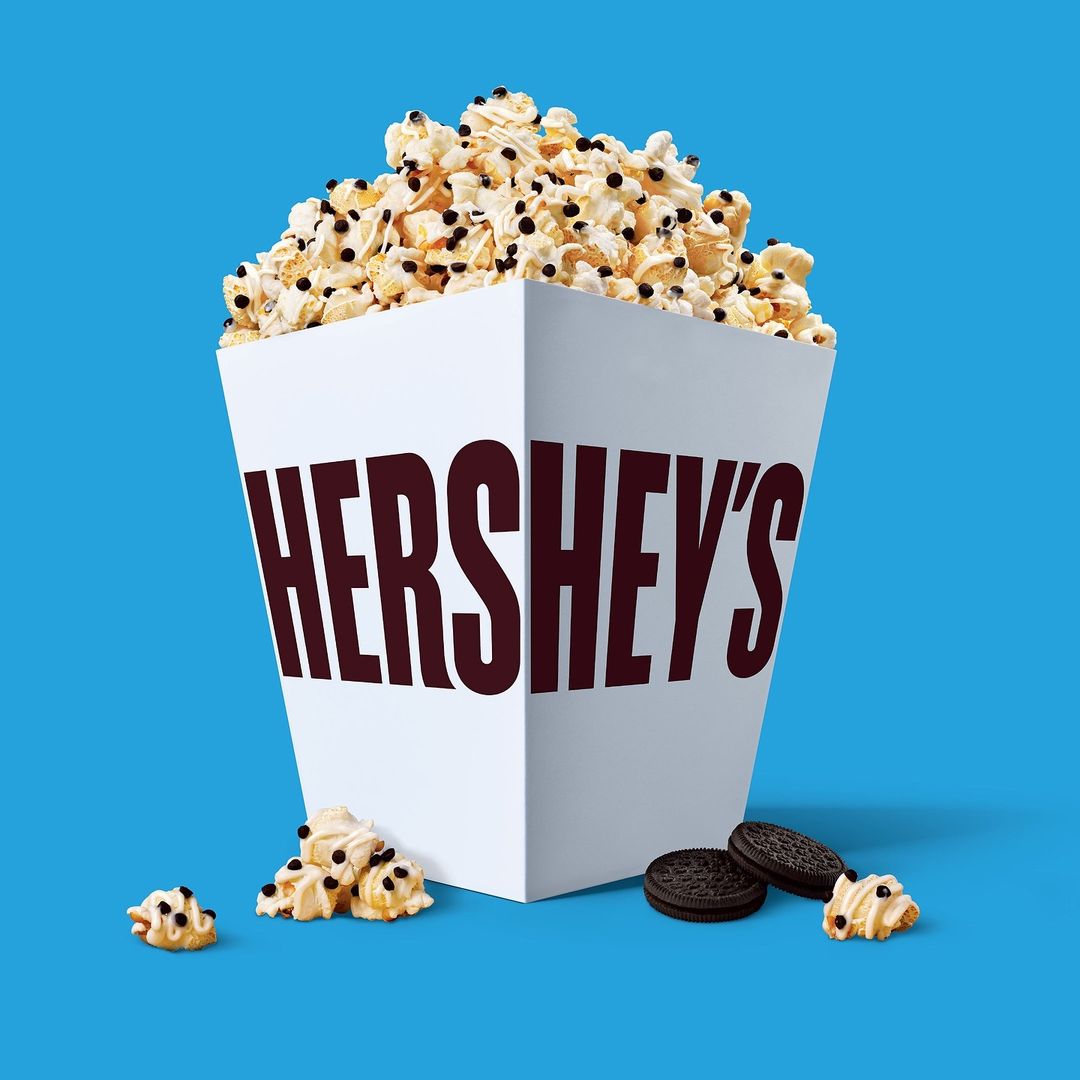 Popcorn overflowing from a white Hershey's branded box, accompanied by cookie pieces, set against a vibrant blue background.