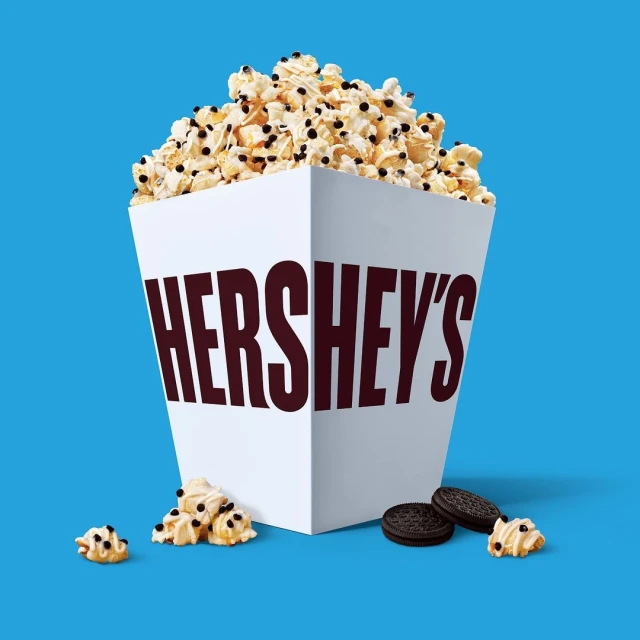Popcorn overflowing from a white Hershey's branded box, accompanied by cookie pieces, set against a vibrant blue background.