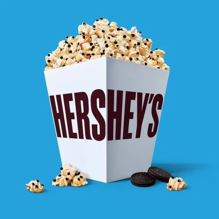 Popcorn overflowing from a white Hershey's branded box, accompanied by cookie pieces, set against a vibrant blue background.