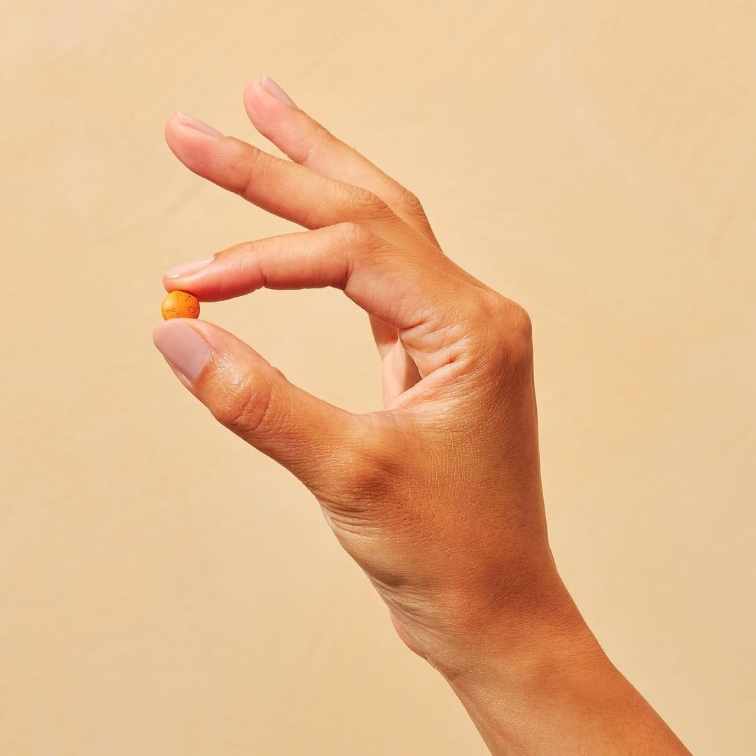 Close-up of an adult human hand pinching a small, round, orange object, against a neutral beige background with soft, diffused lighting.