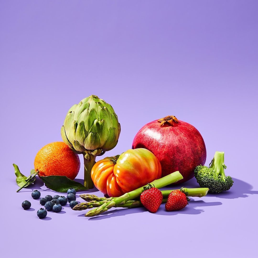 Artichoke, pomegranate, orange, tomato, asparagus, strawberries, blueberries on purple backdrop in a brightly lit food still life.