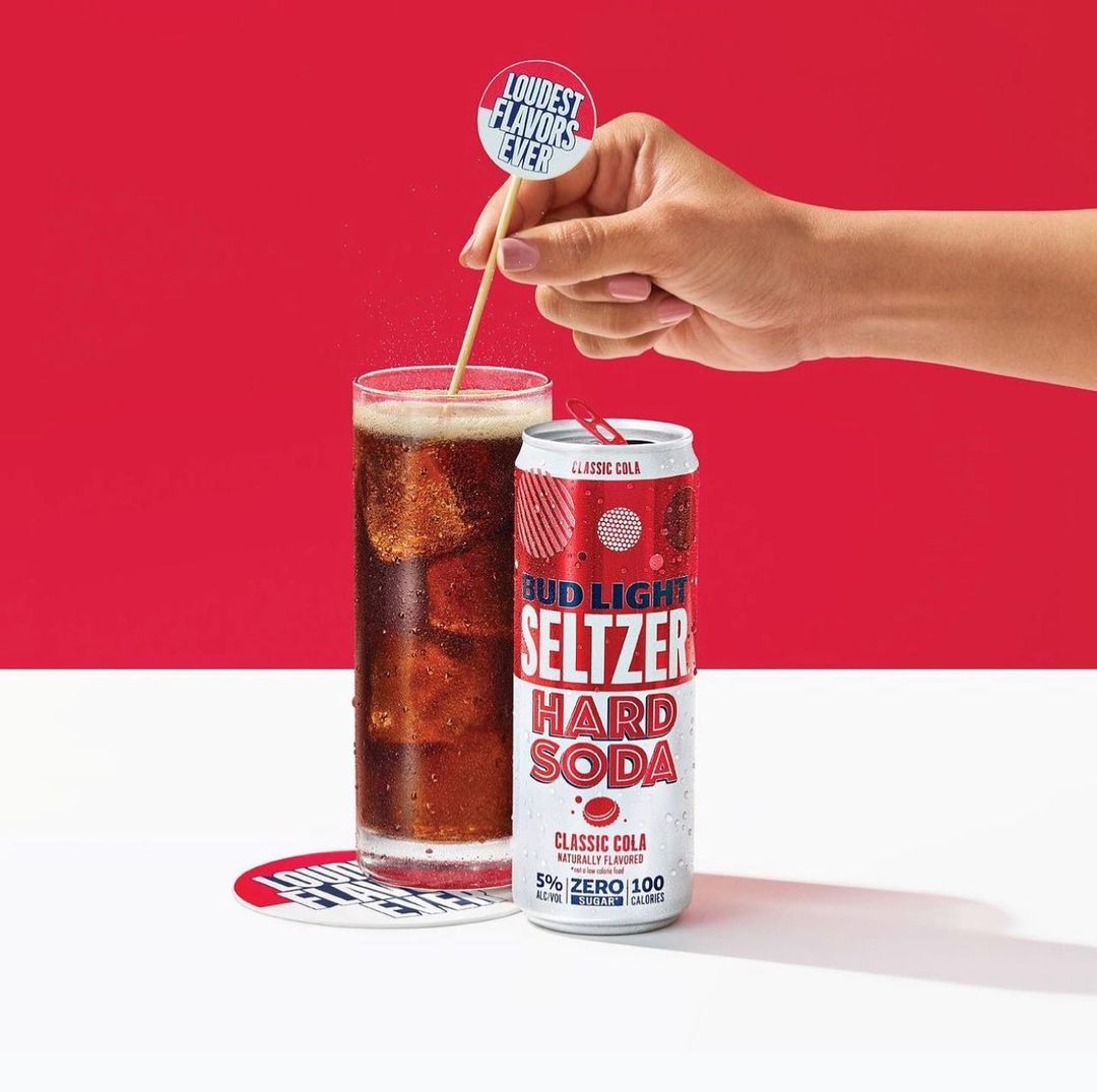 Bud Light Seltzer Hard Soda beverage in a glass with ice and in a can, displayed with a decorative stick, on a white surface against red backdrop.