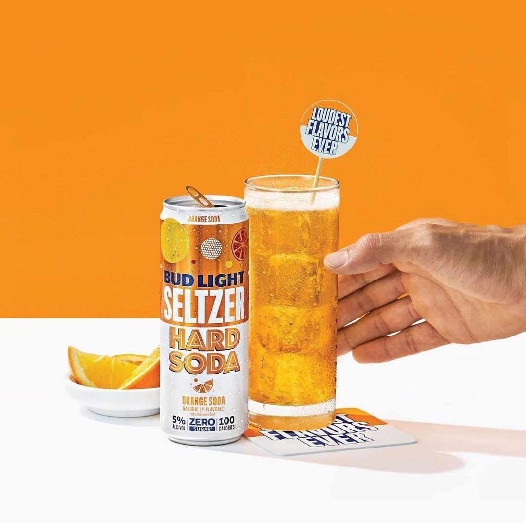 Bud Light Seltzer Hard Soda in can and glass with ice, orange slices, and a hand holding the drink against an orange background.