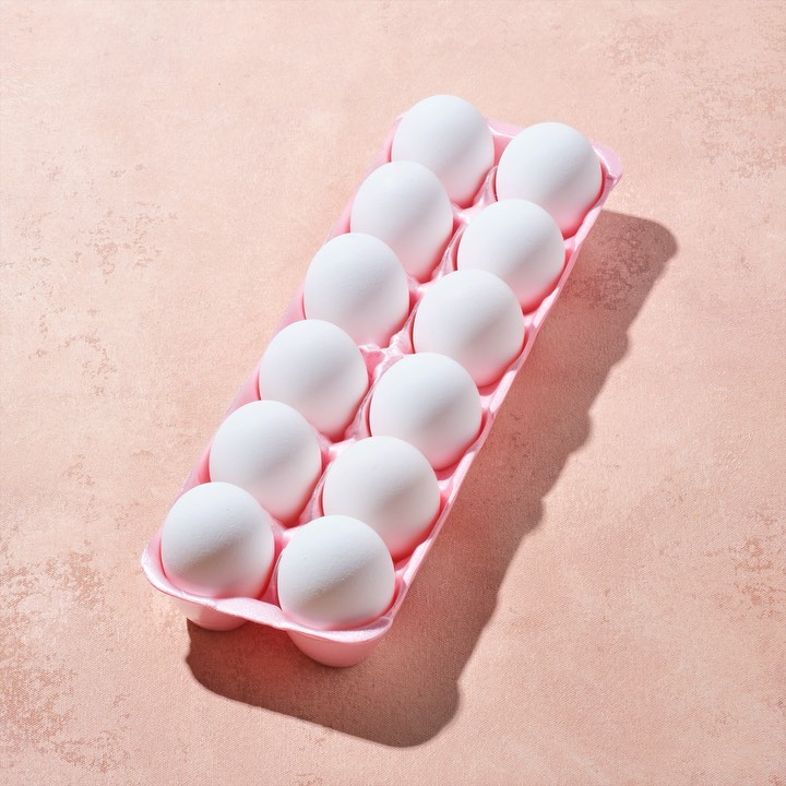Elevated close-up of one dozen white eggs arranged neatly in a pink carton on a peach surface casting shadows, food photography.