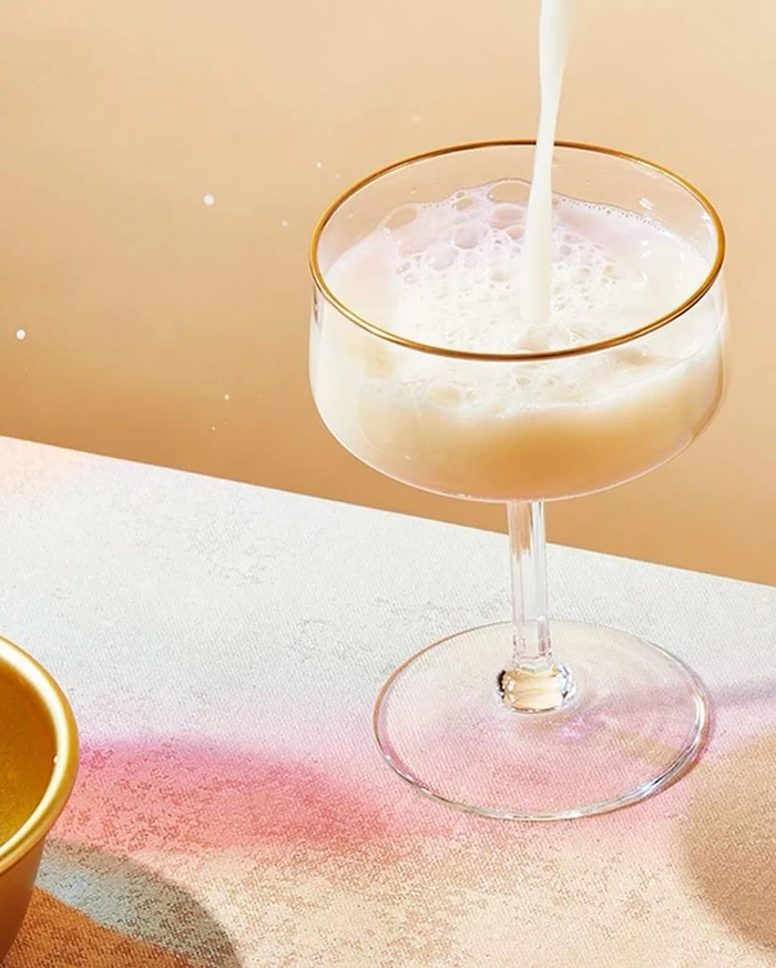 Milk pouring into a stemmed coupe glass with a gold rim on a patterned surface against a warm beige background.