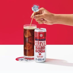 A hand stirring a glass of cola with ice beside a Bud Light Seltzer Hard Soda can against a red and white background
