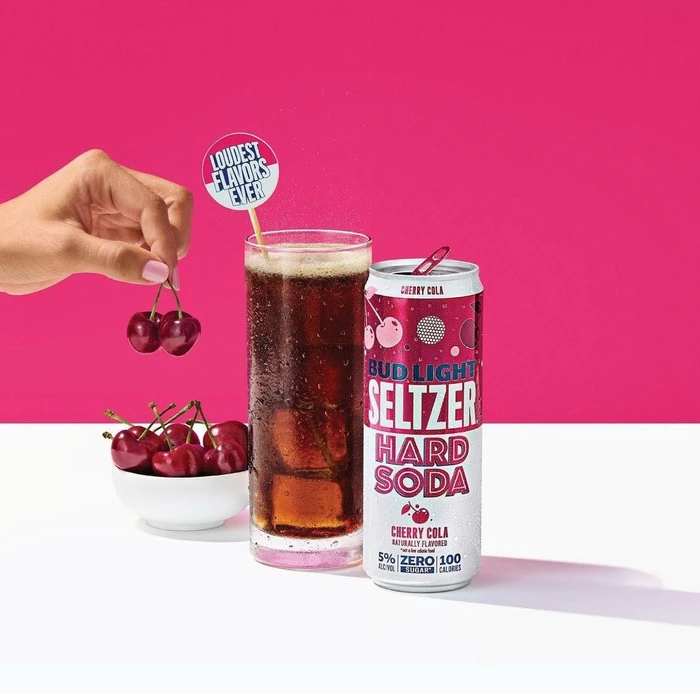 Bud Light Seltzer Hard Soda Cherry Cola can next to a glass of cola with cherries and a hand holding two cherries.