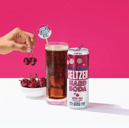 Bud Light Seltzer Hard Soda Cherry Cola can next to a glass of cola with cherries and a hand holding two cherries.
