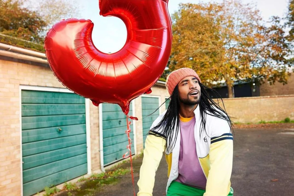 An adult male with dreadlocks holds a large red number balloon outside, wearing a knit cap, lilac shirt, and sporty jacket, smiling.
