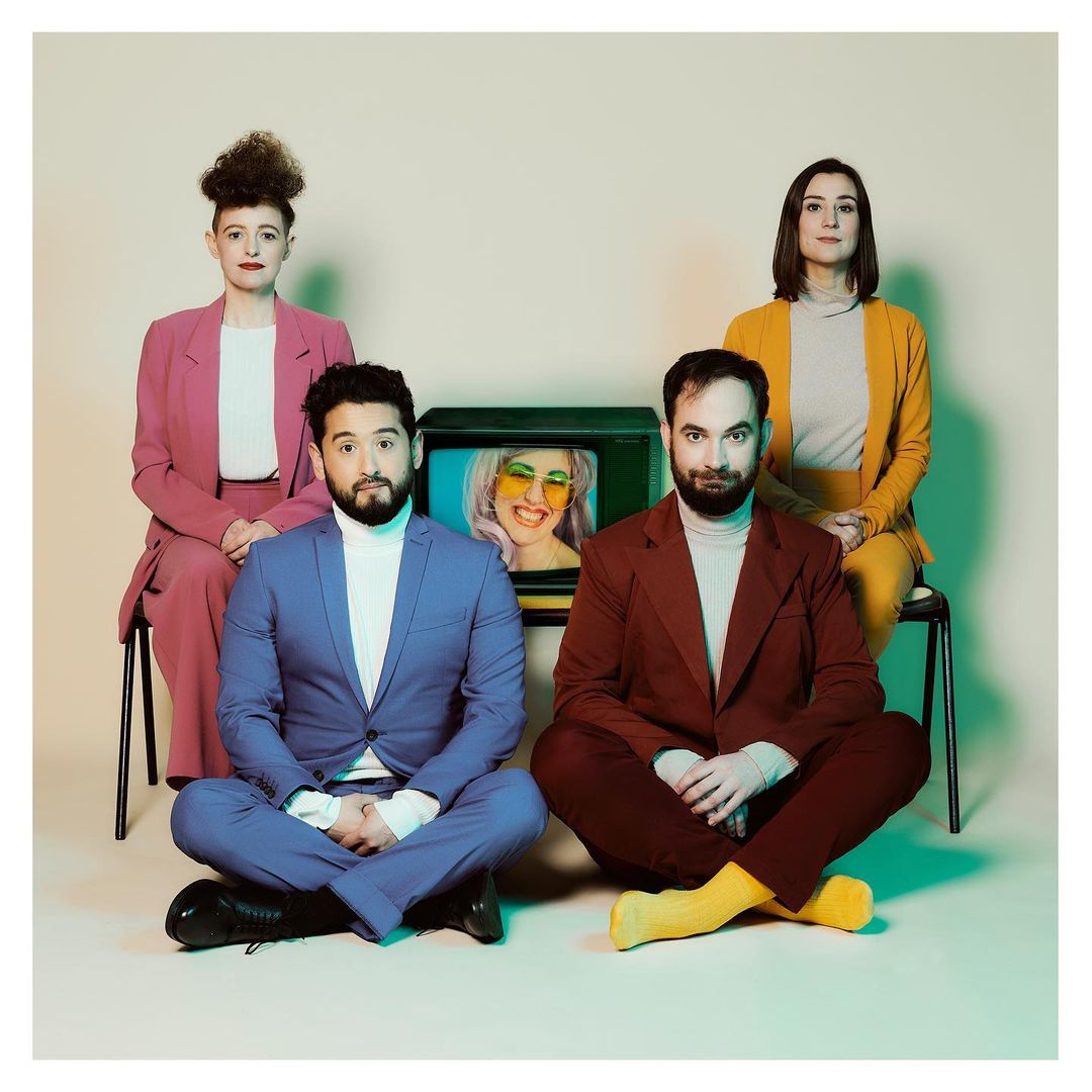 Group portrait of four people in a studio setting; two women seated behind two men in suits, a vintage television set in the middle.