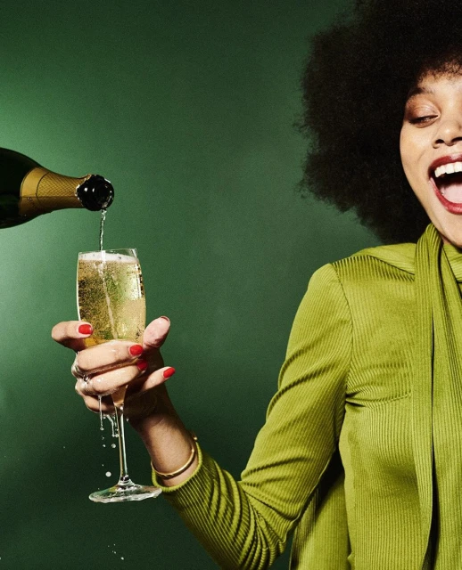 Woman with afro pours champagne into glass against green background. Close-up of celebratory moment, drink, and vintage fashion.