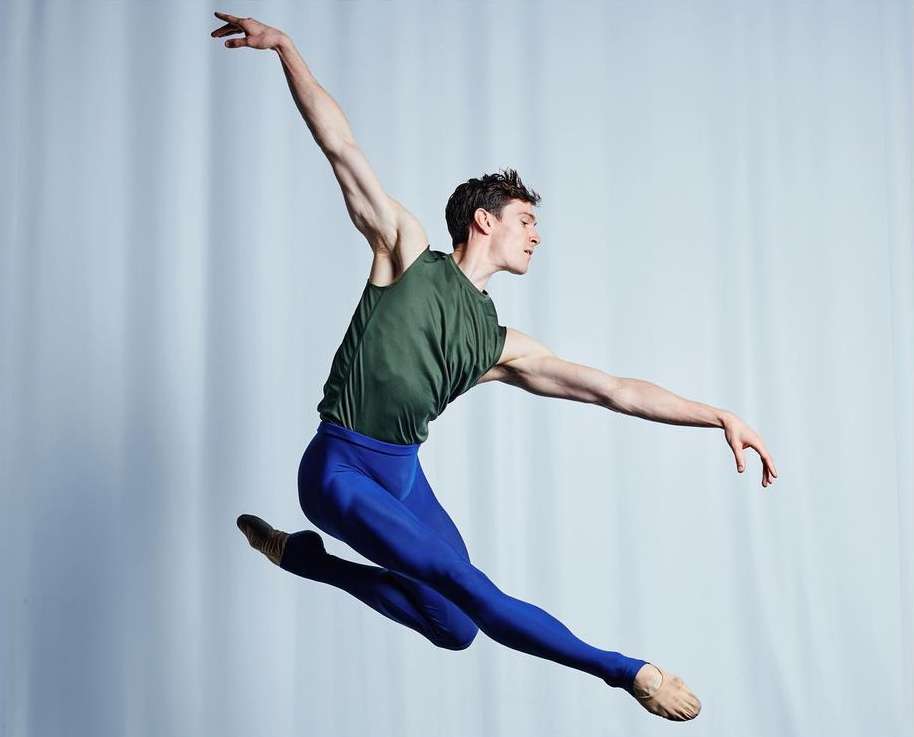 Male ballet dancer in mid-air leap against a soft grey backdrop. Wearing dark green top, blue tights and soft ballet shoes.