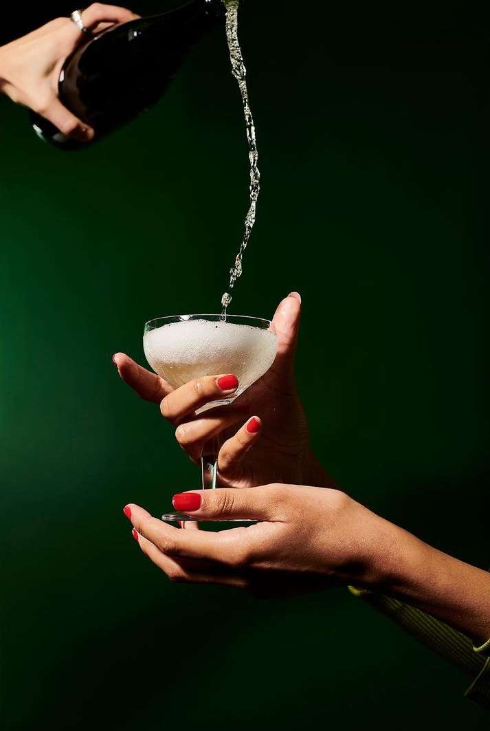 Person pours sparkling beverage into glass held with both hands against a dark green background in studio lighting.