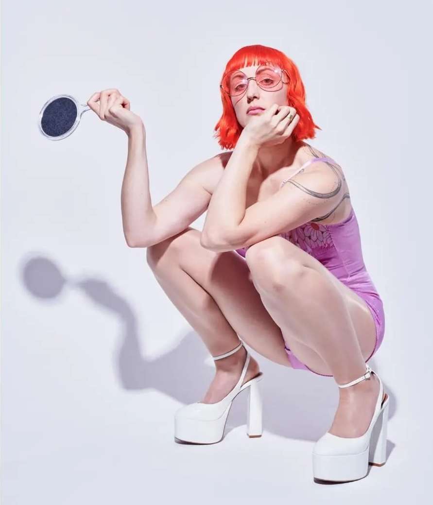 A fair-skinned adult woman with bright orange bob-cut hair, wearing a violet outfit and white platform heels, squats in a white studio.
