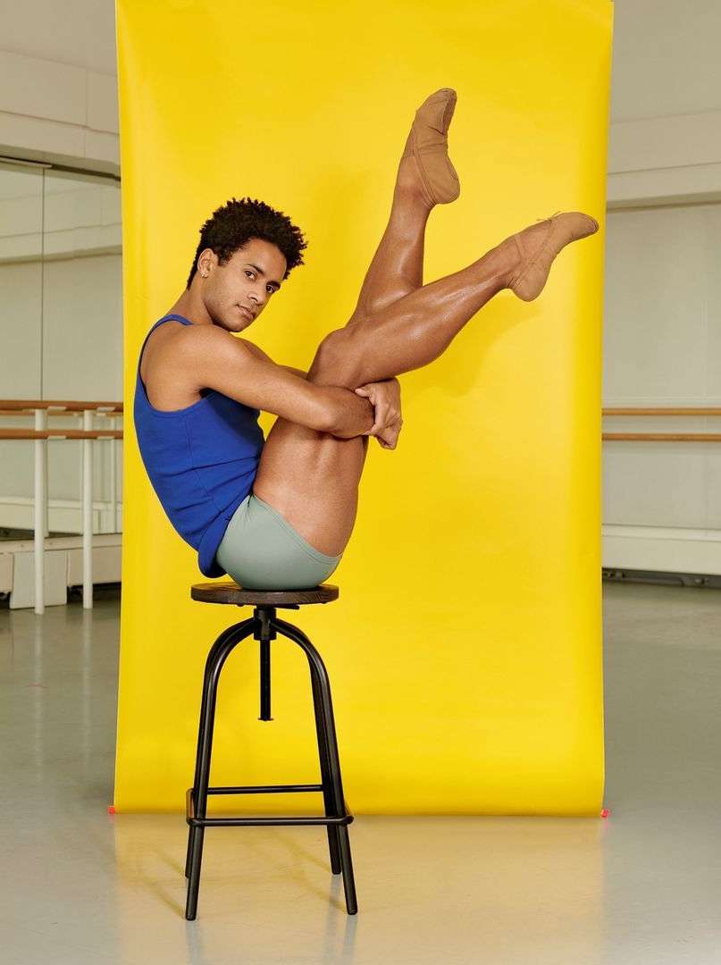 A dancer in blue tank and leotard strikes a pose while seated atop a black stool against a vibrant yellow backdrop in a dance studio.