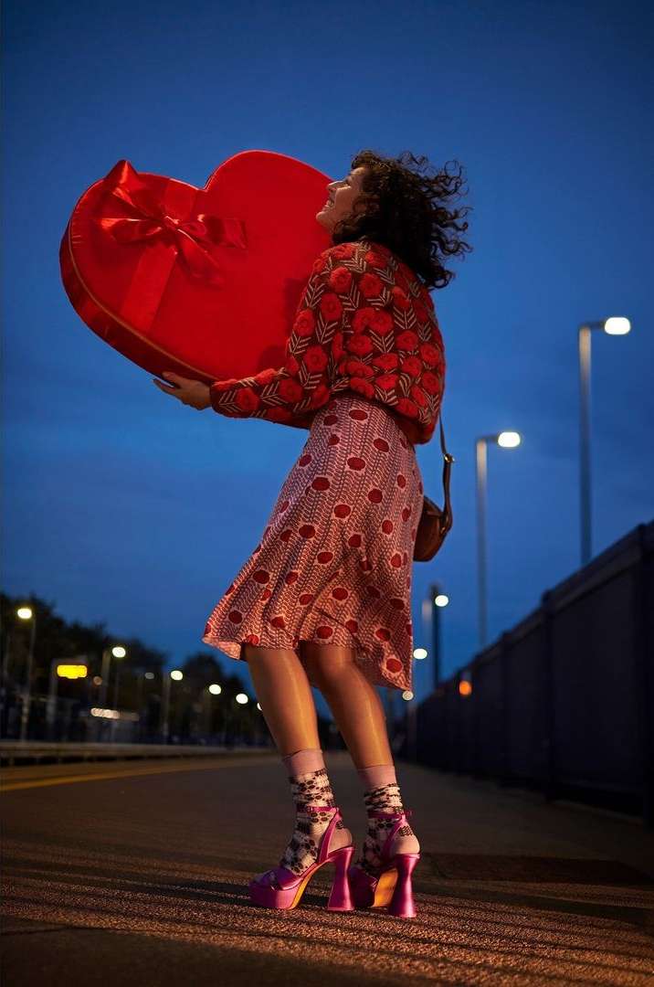 A curly-haired woman in a patterned blouse and skirt carries a large, red, heart-shaped box outdoors under a dark blue sky.