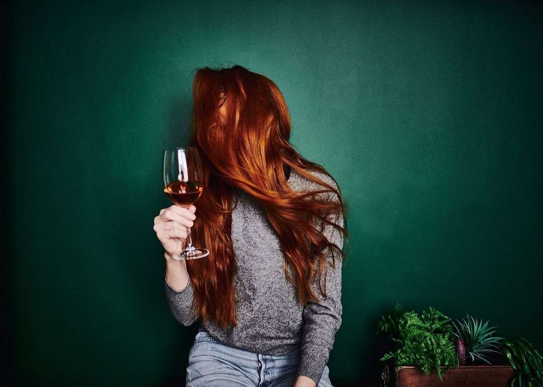 A woman with long red hair holding a wine glass against a dark green background, wearing a gray sweater and jeans.